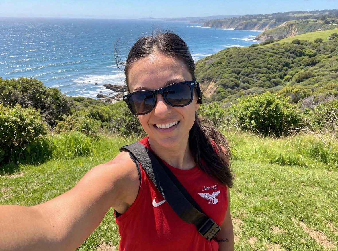 Romi smiling on a coastal hike, with the ocean behind her