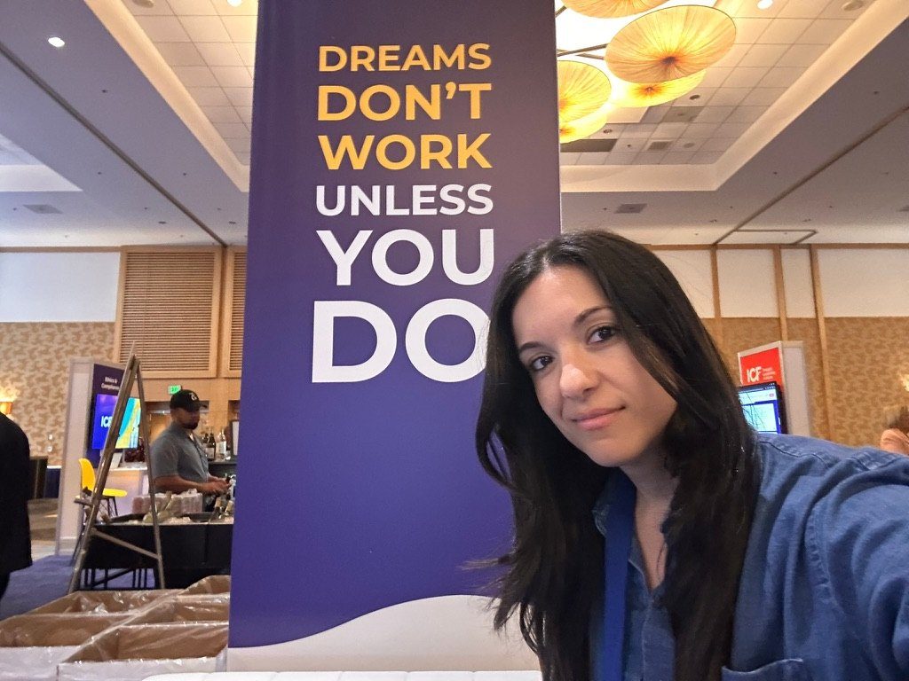 Romi at an International Coaching Federation event in Paris, beside a banner that reads Dreams don't work unless you do