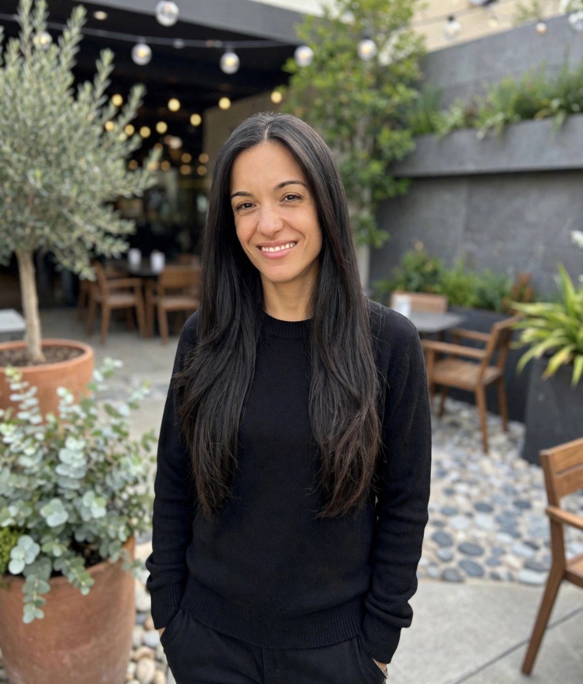 Romi smiling in a black sweater on a garden patio