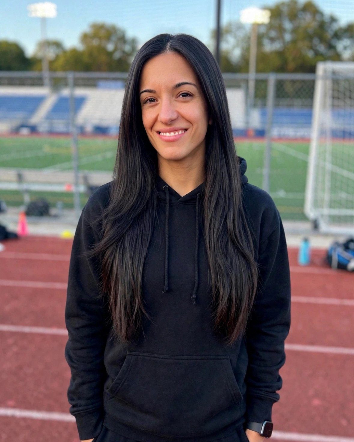 Romi smiling in a black hoodie on a running track beside a soccer field