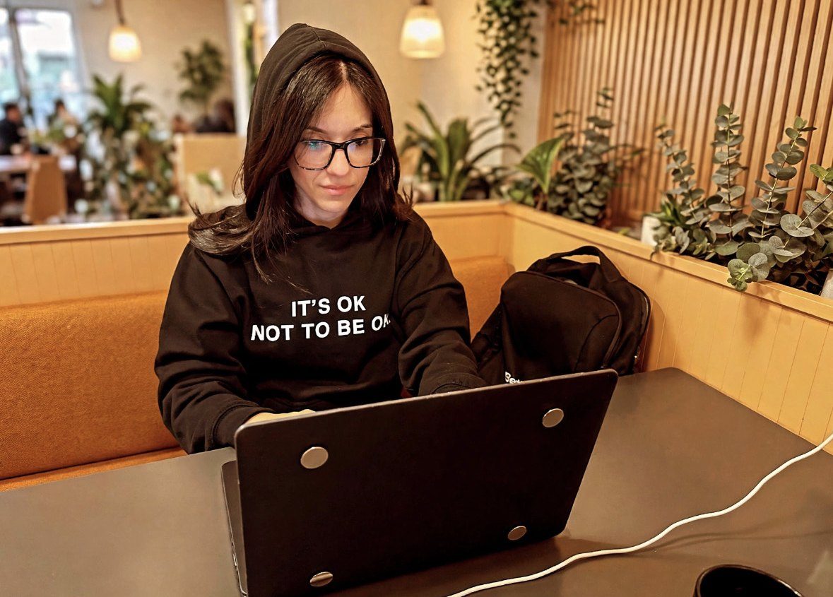 Romi at her laptop for a video session, in a hoodie that says It's OK not to be OK